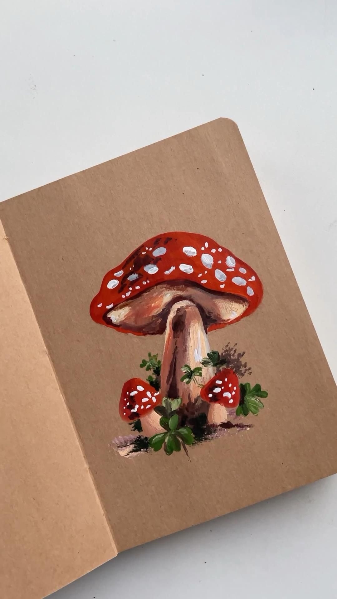 Cây nấm mũ đỏ chấm trắng vẽ gouache trên sổ giấy kraft, có hai nấm nhỏ và lá xanh dưới gốc