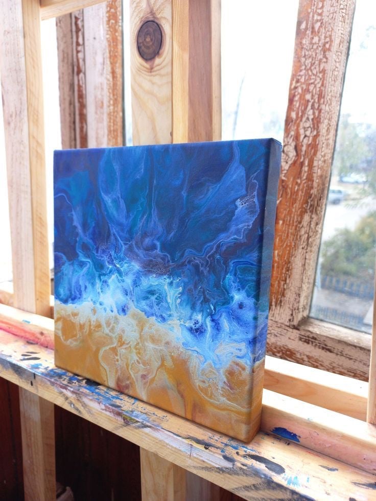 Tranh fluid art khổ vuông nhỏ của Eva Anastasia đặt bên cửa sổ gỗ: màu xanh dương đậm loang xuống mảng vàng cát như sóng biển vỗ bờ