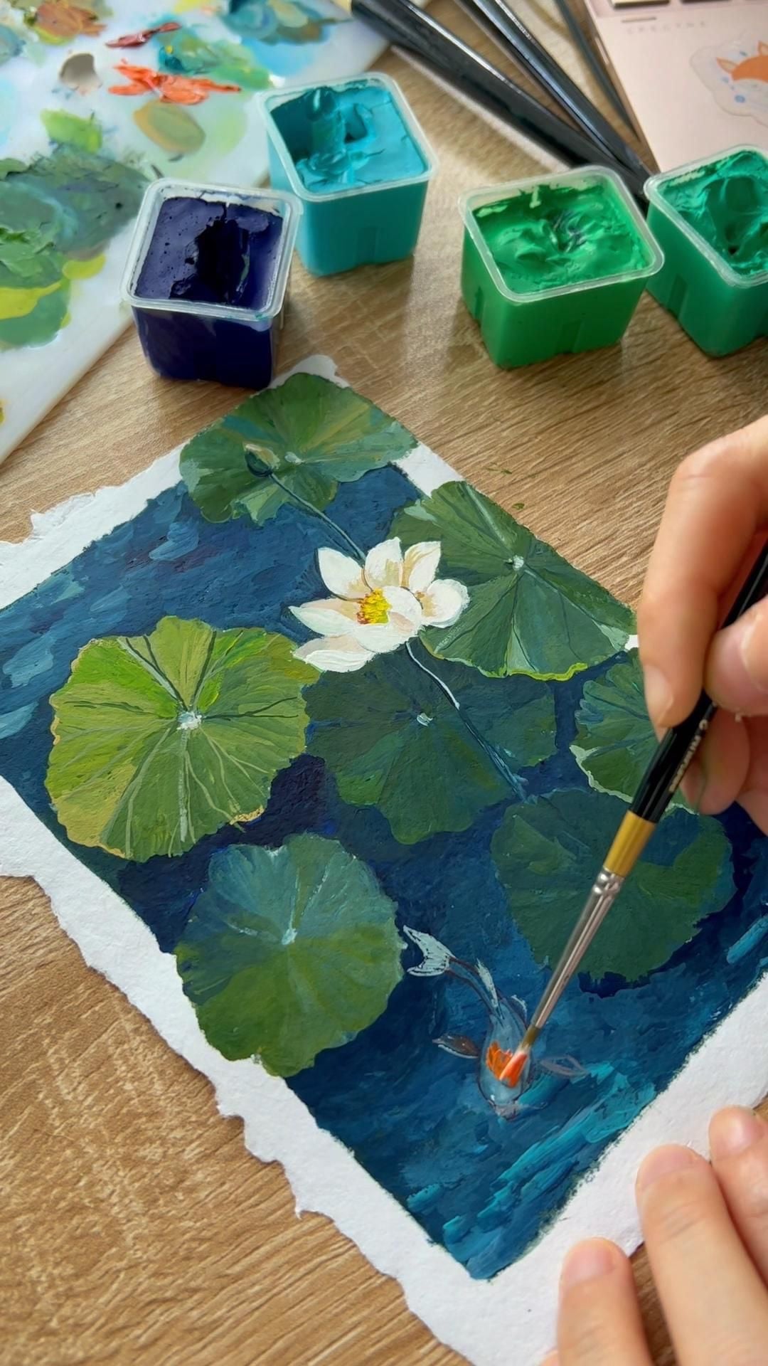 Bàn vẽ gouache: các hũ màu xanh, cọ, palette và bức tranh lá sen cá koi đang vẽ dở