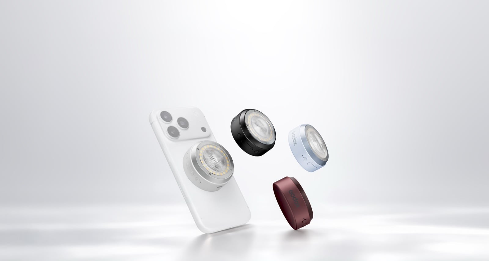Một chiếc iPhone màu bạc nhìn từ mặt lưng, bên cạnh là ba chiếc đèn Godox A2 hình tròn màu bạc, xanh nhạt và đỏ sẫm lơ lửng trên nền trắng