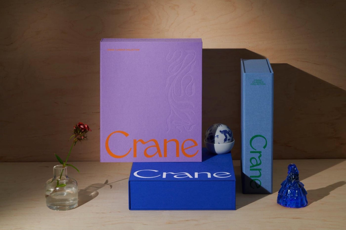 Các hộp giấy Crane màu tím, xanh dương và xanh nhạt xếp đứng trên mặt bàn gỗ, cạnh một bình hoa nhỏ và tượng gốm xanh trắng, chữ Crane màu cam đỏ