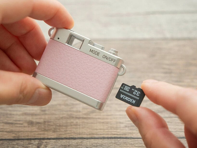 Hai tay cầm chiếc máy ảnh Pieni III màu hồng phấn, tay kia cầm thẻ nhớ microSD 32GB đặt cạnh khe thẻ của máy, nền gỗ