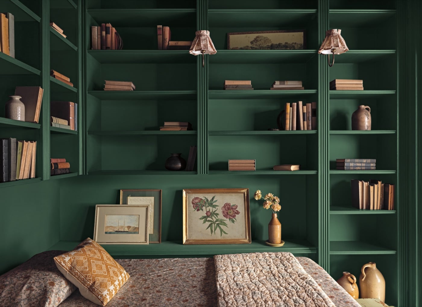 Phòng đọc sách với hệ kệ âm tường và tường sơn xanh lá trầm Webster Green, giường nghỉ phủ chăn len, gối và tranh hoa đặt trên kệ