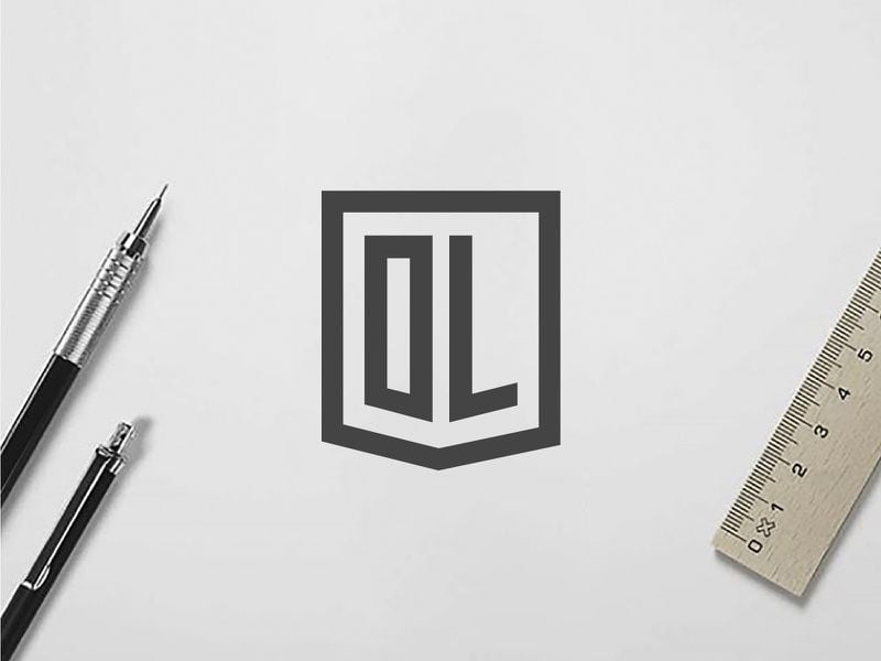 Logo DL nét xám đậm trong khung khiên, đặt trên giấy cạnh bút chì và thước kẻ