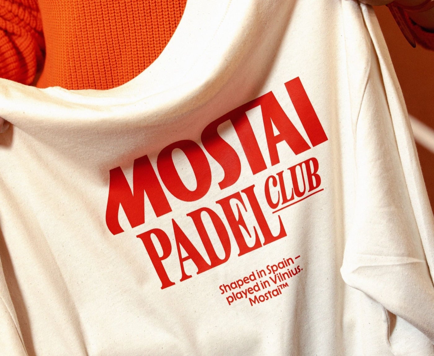 Áo phông màu kem in chữ MOSTAI PADEL CLUB màu đỏ và dòng chữ nhỏ Shaped in Spain, played in Vilnius, phía sau là áo len đan màu cam