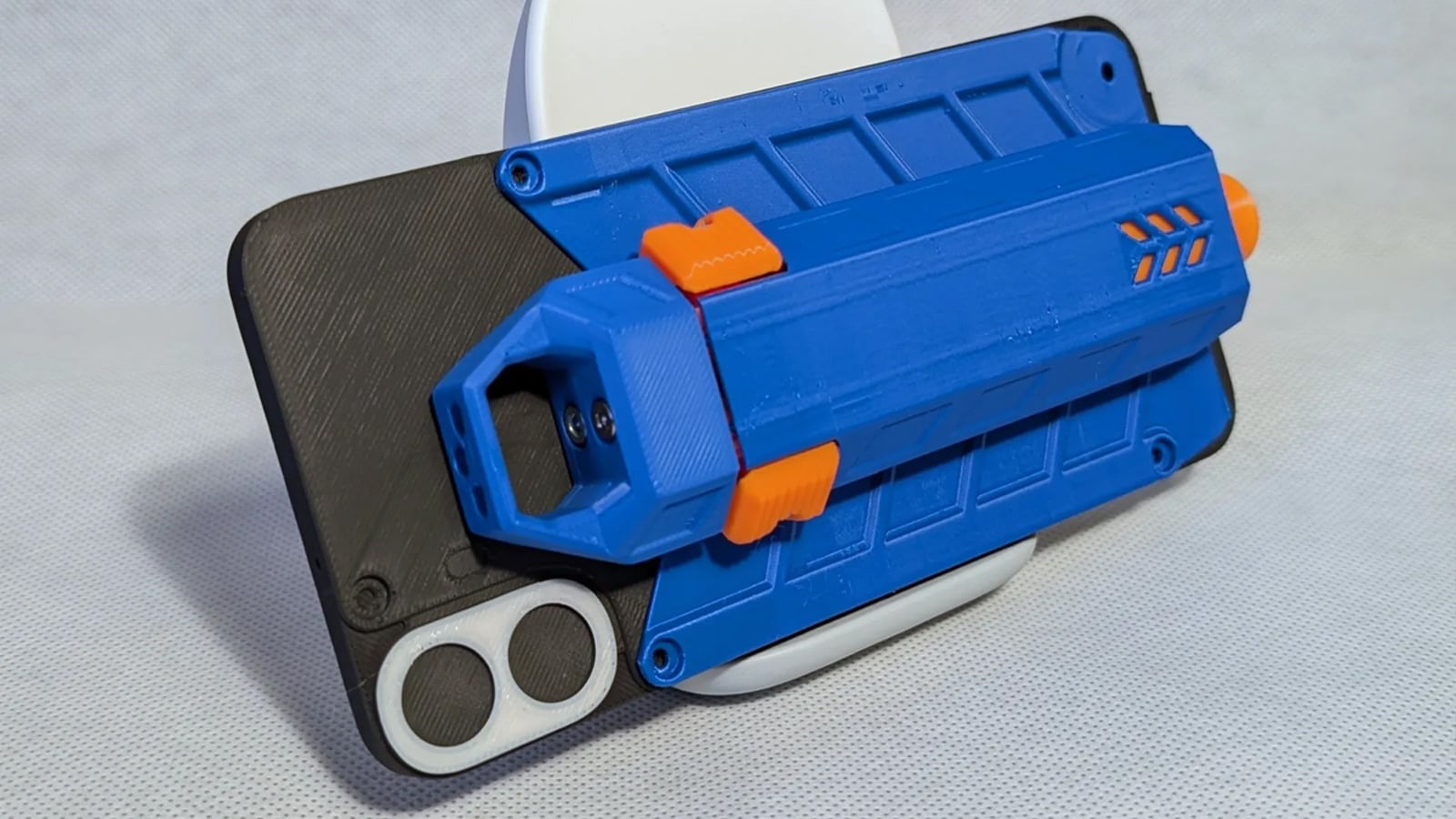 Mô-đun bắn đạn xốp in 3D màu xanh dương và cam gắn vào mặt lưng một chiếc điện thoại đặt nghiêng trên bàn
