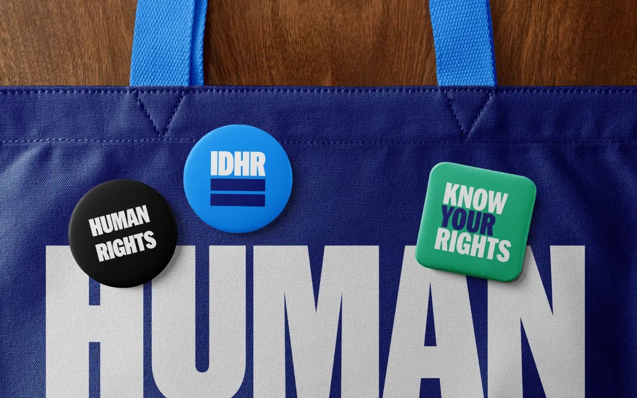 Túi vải xanh dương in chữ HUMAN trắng rất lớn, trên túi gắn ba huy hiệu tròn Human Rights, IDHR và Know Your Rights